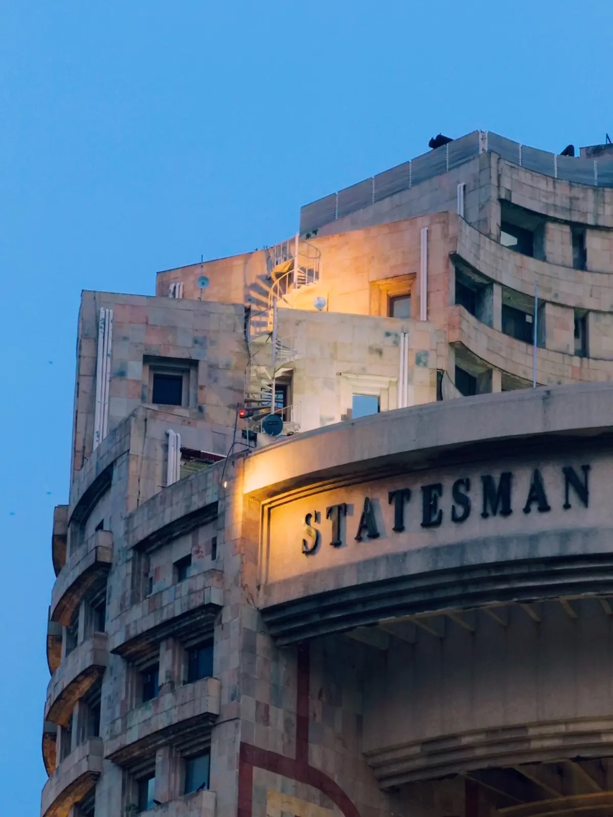 Warm lights on the curved Statesman building beneath a deep blue evening sky