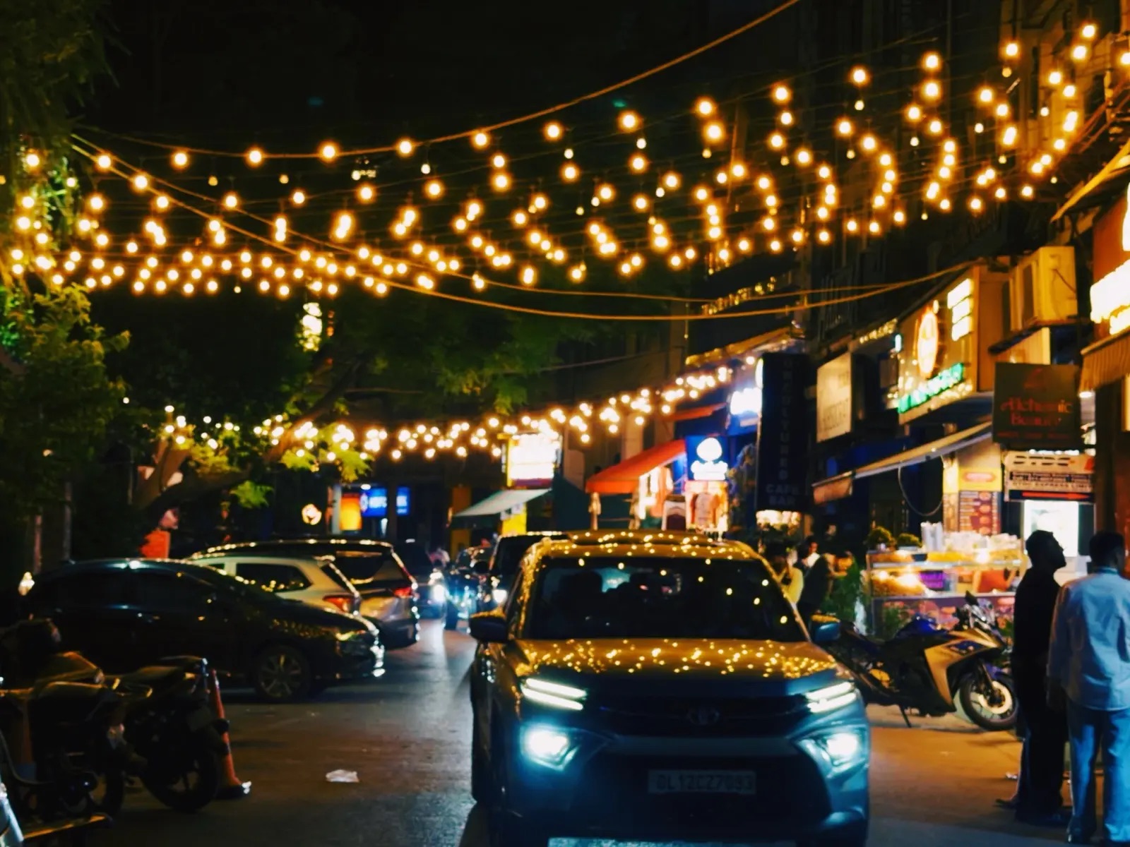 An evening street strung with warm lights, with cars waiting in line